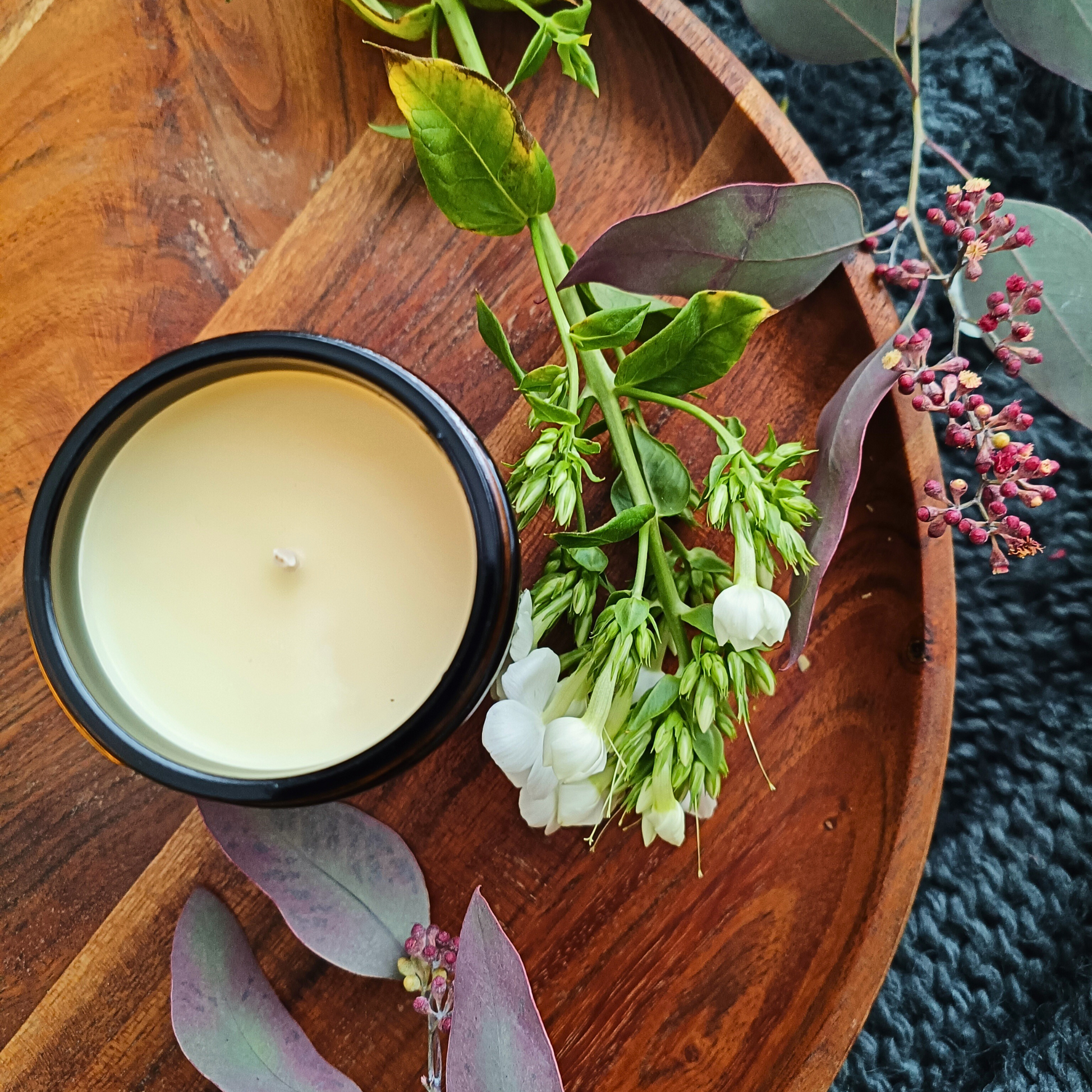 An open candle in an amber glass jar sitting on a brown wooden tray with some a flower stem