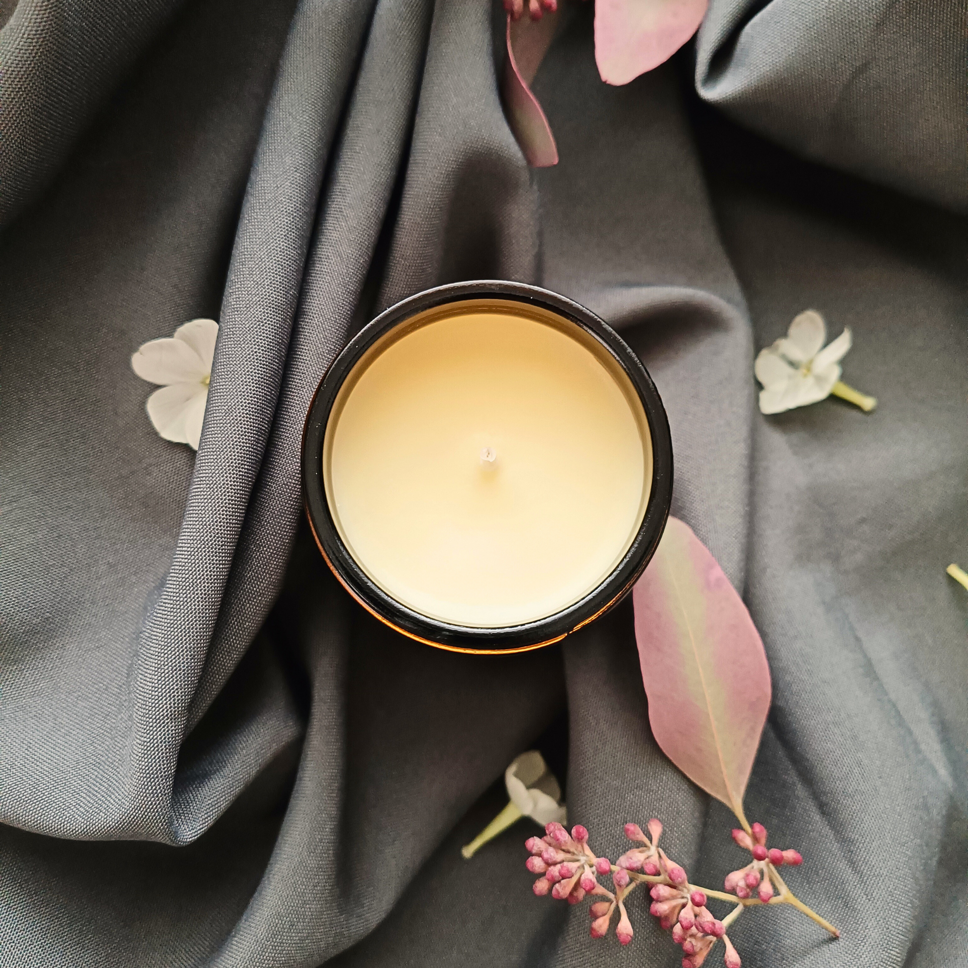 An open candle jar resting on a grey sheet with scattered flowers
