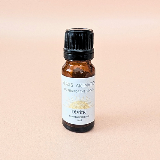 A 10ml amber glass bottle of Nicky's Aromatics 'Divine' essential oil blend made with orange, frangipani and patchouli. With a black cap and white label, set against a solid pale peach background.