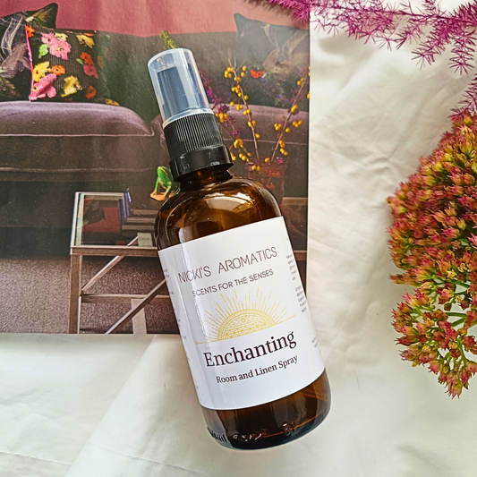 A 100ml amber glass bottle of Enchanting room and linen spray resting on a white sheet and magazine
