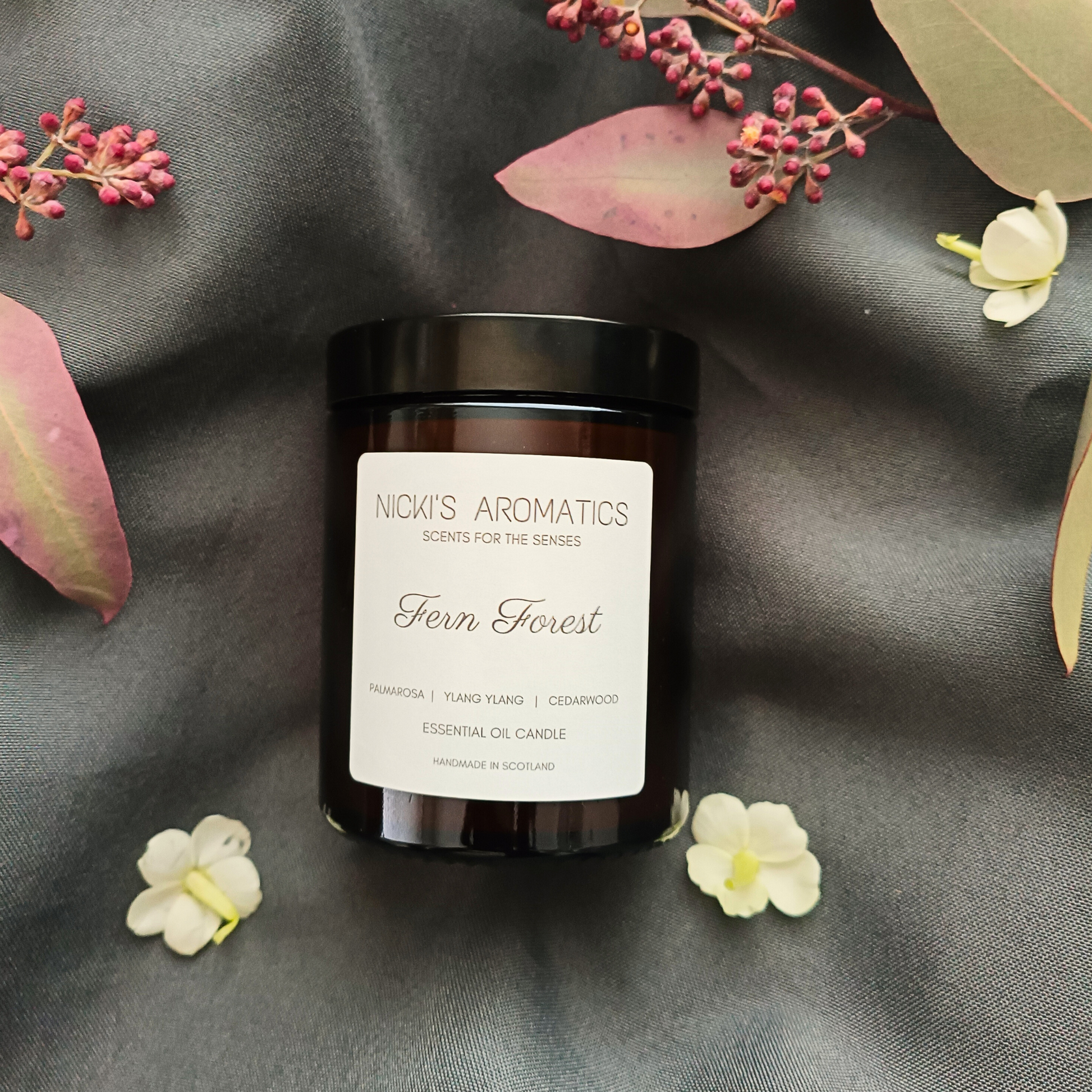 A Fern Forest candle in an amber glass jar resting on a grey sheet with leaves and flowers
