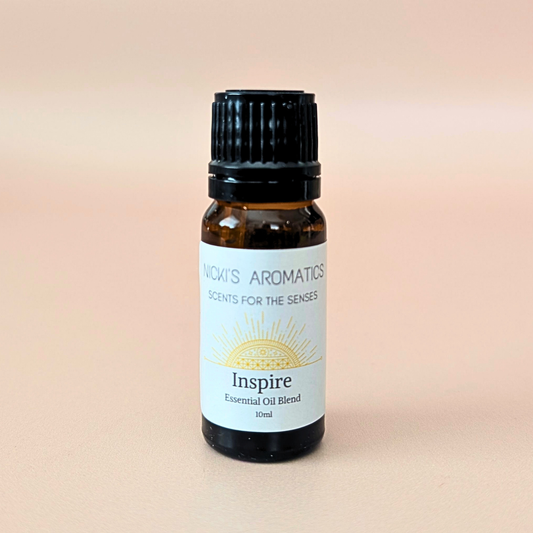 A 10ml amber glass bottle of 'Inspire' essential oil blend by 'Nicki's Aromatics' made with eucalyptus, bay and clove. With a black ribbed cap and a white label featuring a gold sun graphic, set against a soft peach background.