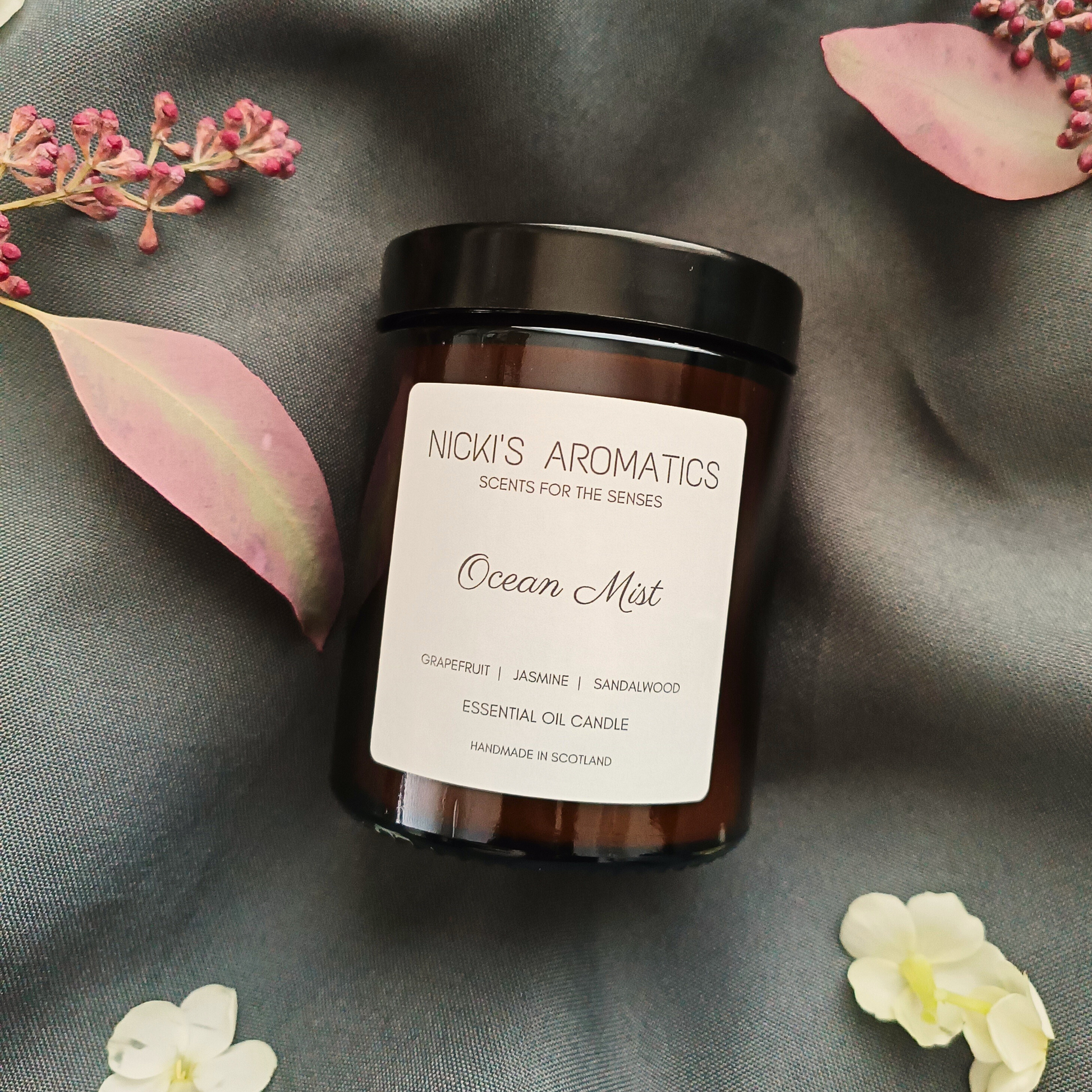 An Ocean Mist essential oil candle in an amber glass jar resting on a grey sheet with some flowers and leaves