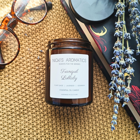 Natural essential oil candle in an amber glass jar resting on a throw near a pair of glasses, a book and some lavender stems