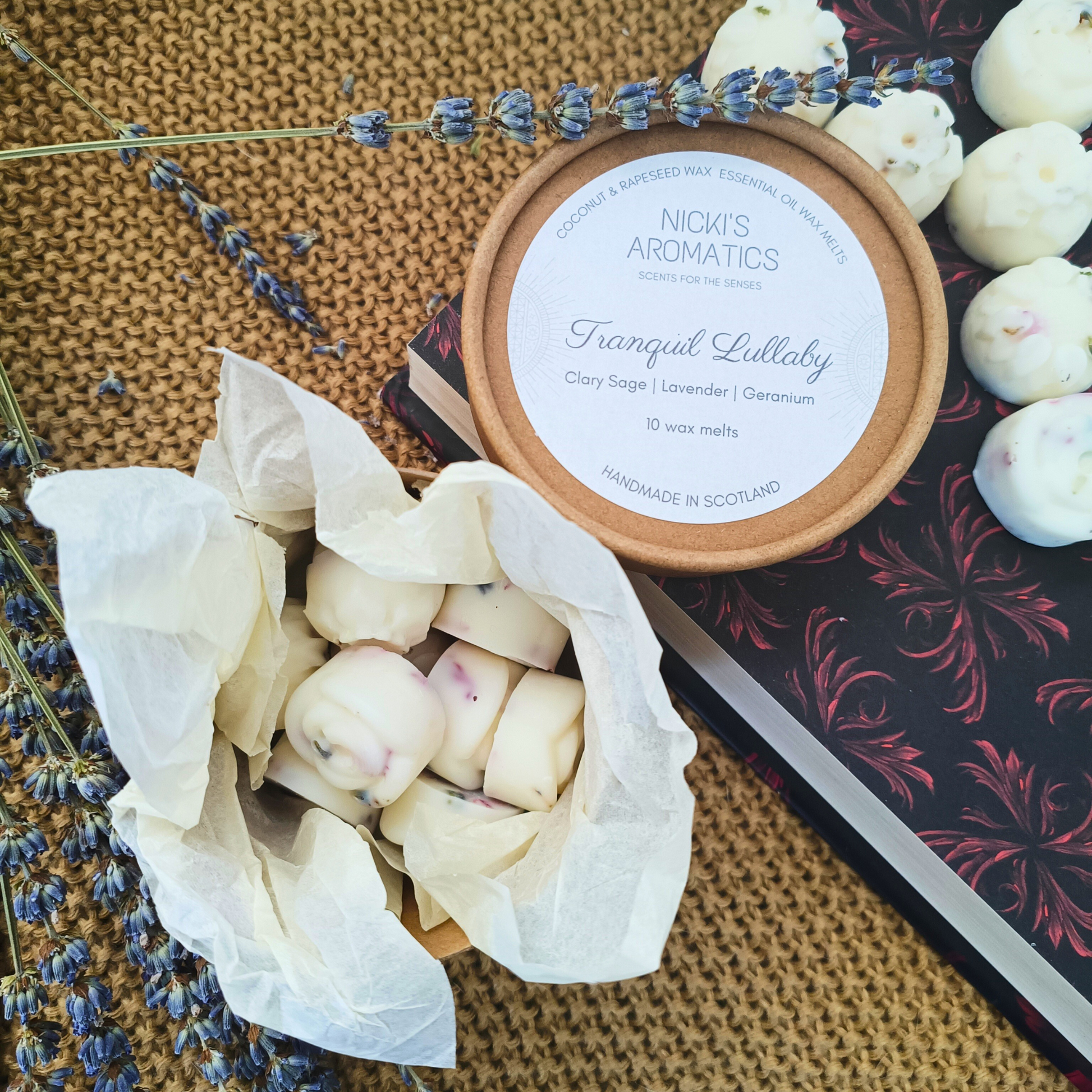 An open kraft tub containing 10 individual wax melts sat on tissue paper which is surrounded by lavender stalks