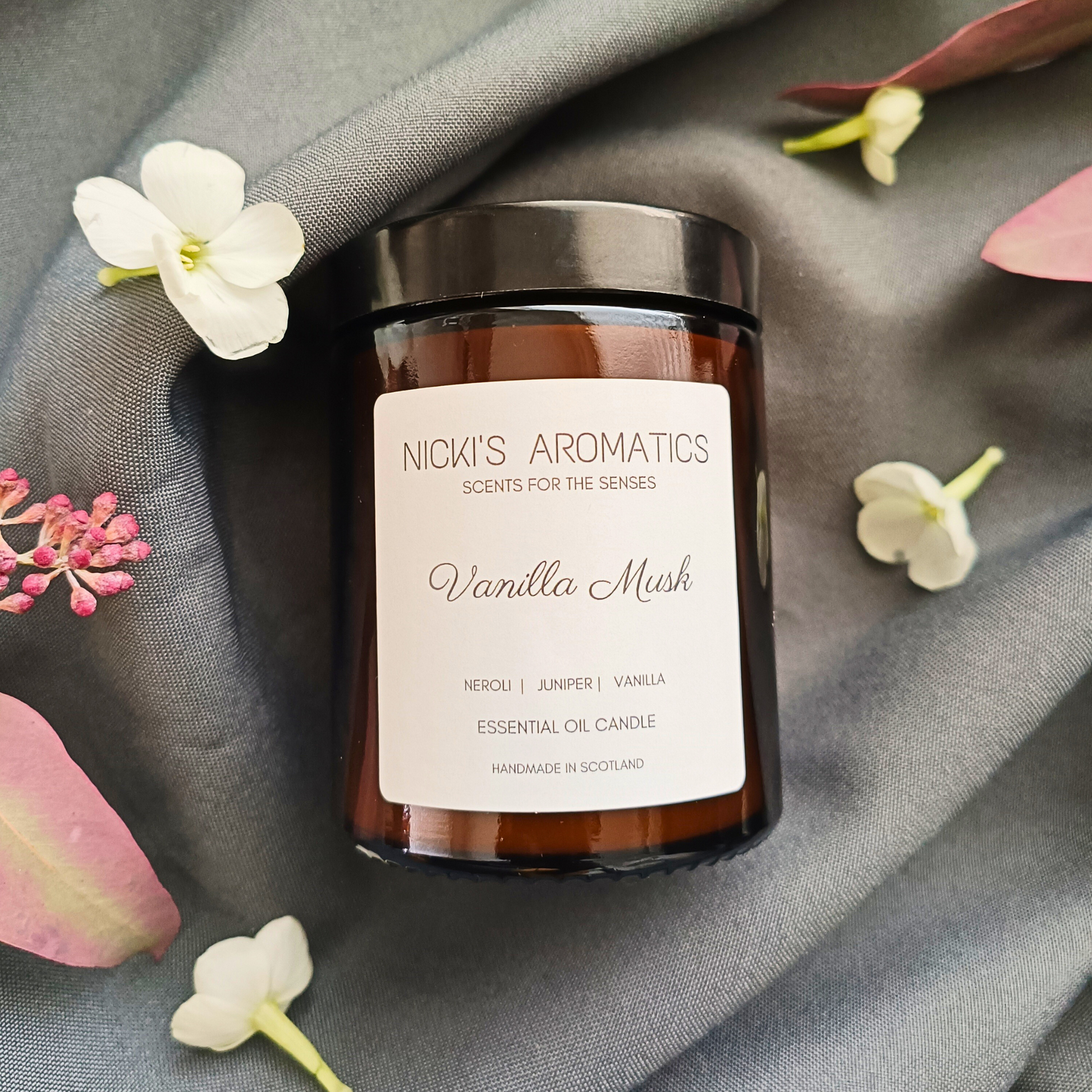 A Vanilla Musk essential oil candle in an amber glass jar resting on a grey sheet surrounded by flowers