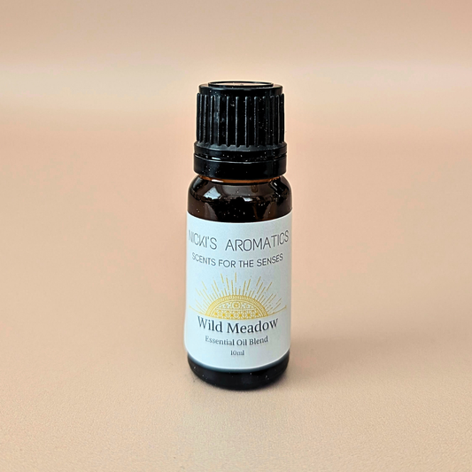 A small amber glass bottle of NICKI'S AROMATICS 'Wild Meadow' essential oil blend made with orange, ylang-ylang and cedarwood, with a black ribbed cap and a white label, set against a solid beige background.