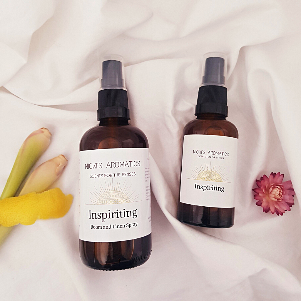 A 100ml and 50ml amber glass bottles of Inspiriting essential oil room and linen spray resting on a white sheet with a rind of lemon and lemongrass