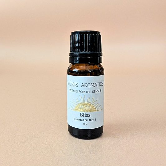 A 10ml amber glass bottle of Nicki's Aromatics 'Bliss' essential oil blend with bergamot, ylang-ylang and patchouli. With a black cap and white label, set against a soft peach background.