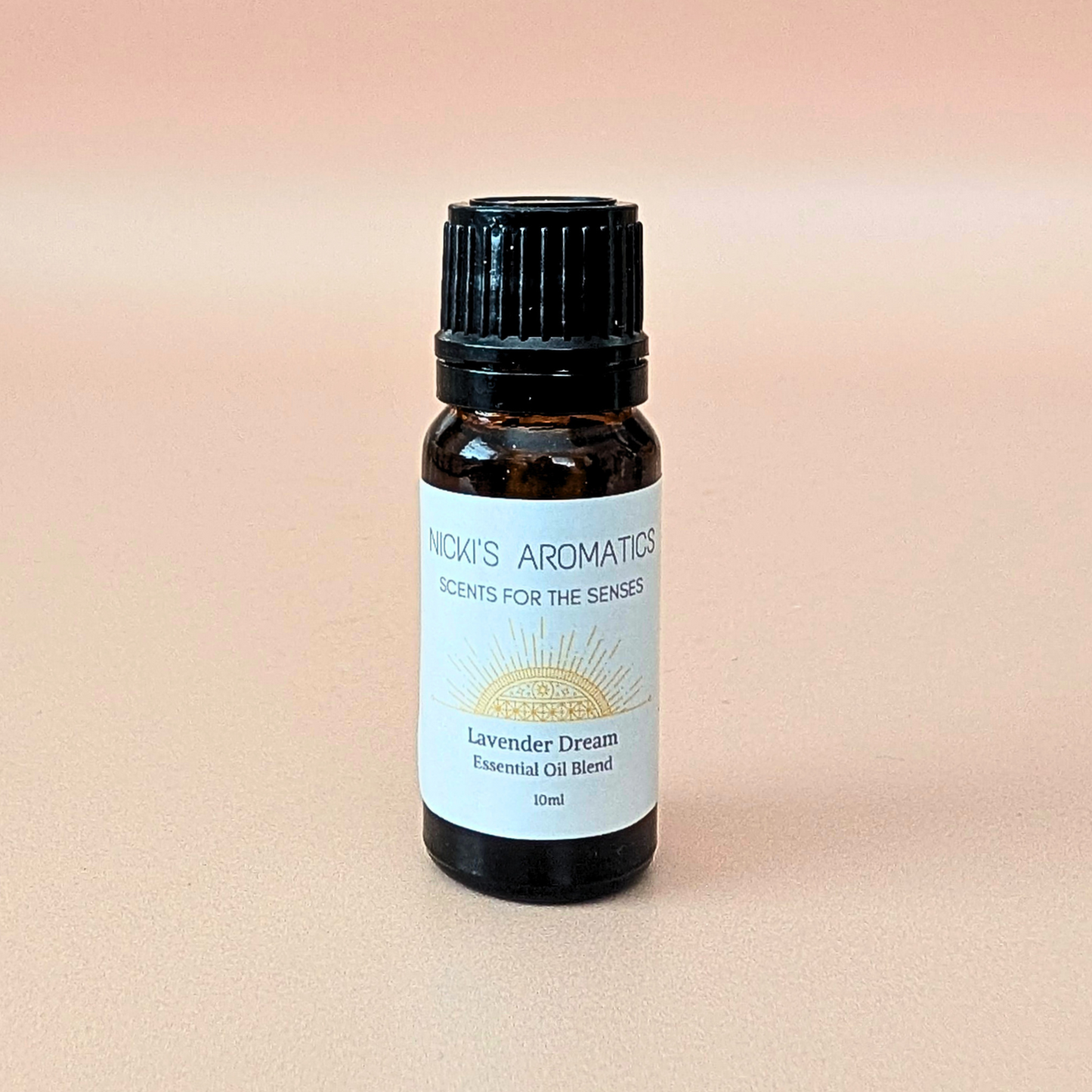 A small amber glass bottle of Lavender Dream essential oil blend made with lavender, chamomile and bergamot. With a black ribbed cap and a white label reading 'Nicki's Aromatics' and 'Lavender Dream', centred on a soft peach background.