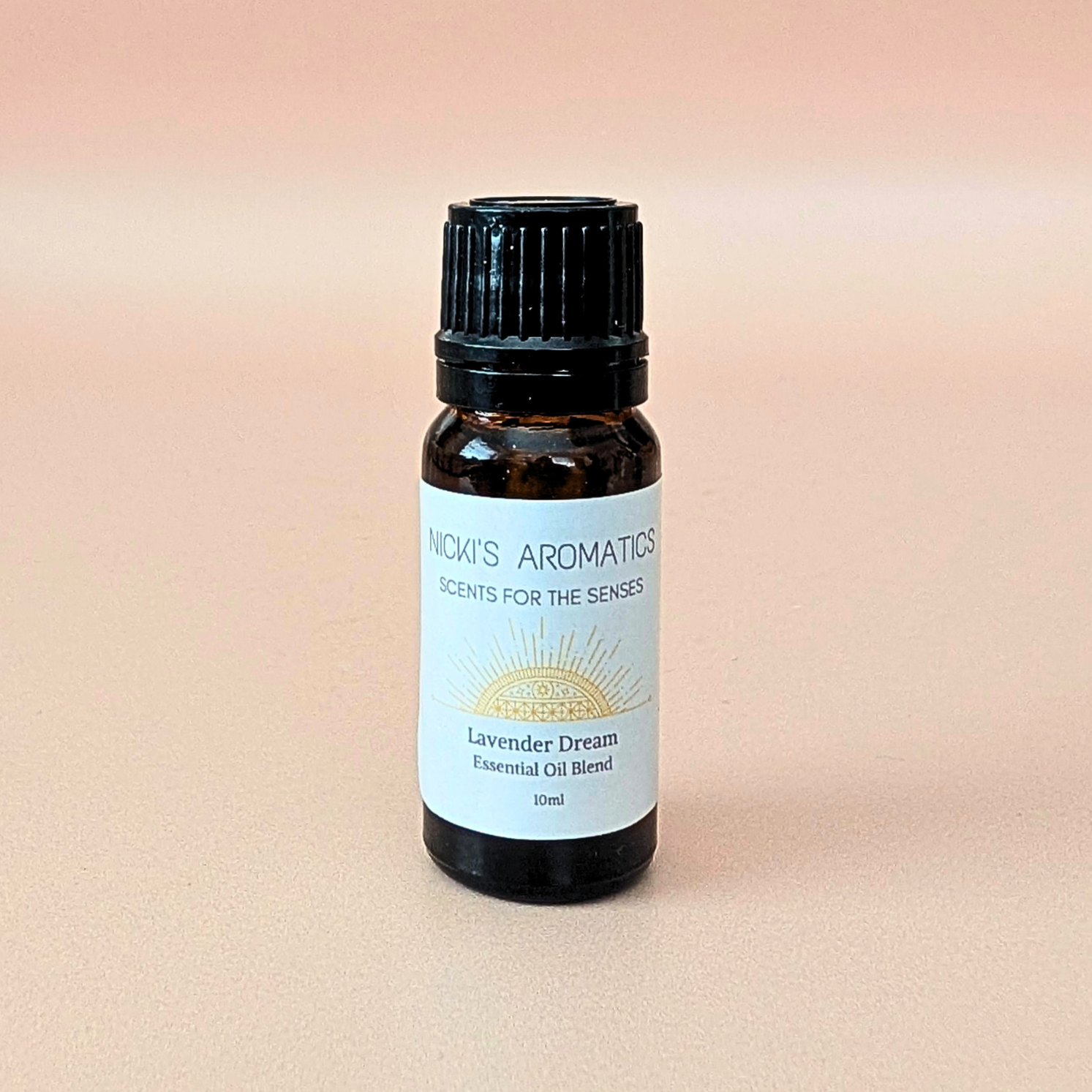 A small amber glass bottle of Lavender Dream essential oil blend made with lavender, chamomile and bergamot. With a black ribbed cap and a white label reading 'Nicki's Aromatics' and 'Lavender Dream', centred on a soft peach background.
