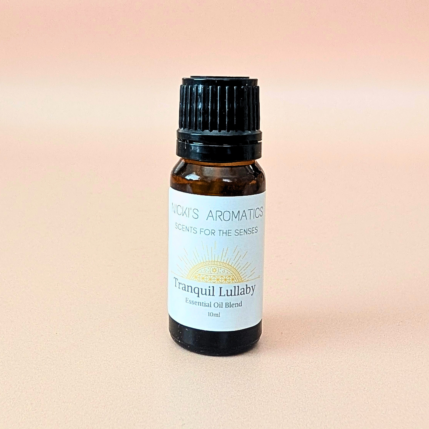 A small amber glass bottle of essential oil blend made with clary sage, lavender and geranium, with a black ribbed cap and a white label reading 'Nicki's Aromatics' and 'Tranquil Lullaby', centered on a soft peach-pink background.