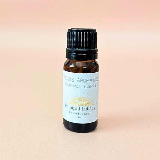 A small amber glass bottle of essential oil blend made with clary sage, lavender and geranium, with a black ribbed cap and a white label reading 'Nicki's Aromatics' and 'Tranquil Lullaby', centered on a soft peach-pink background.