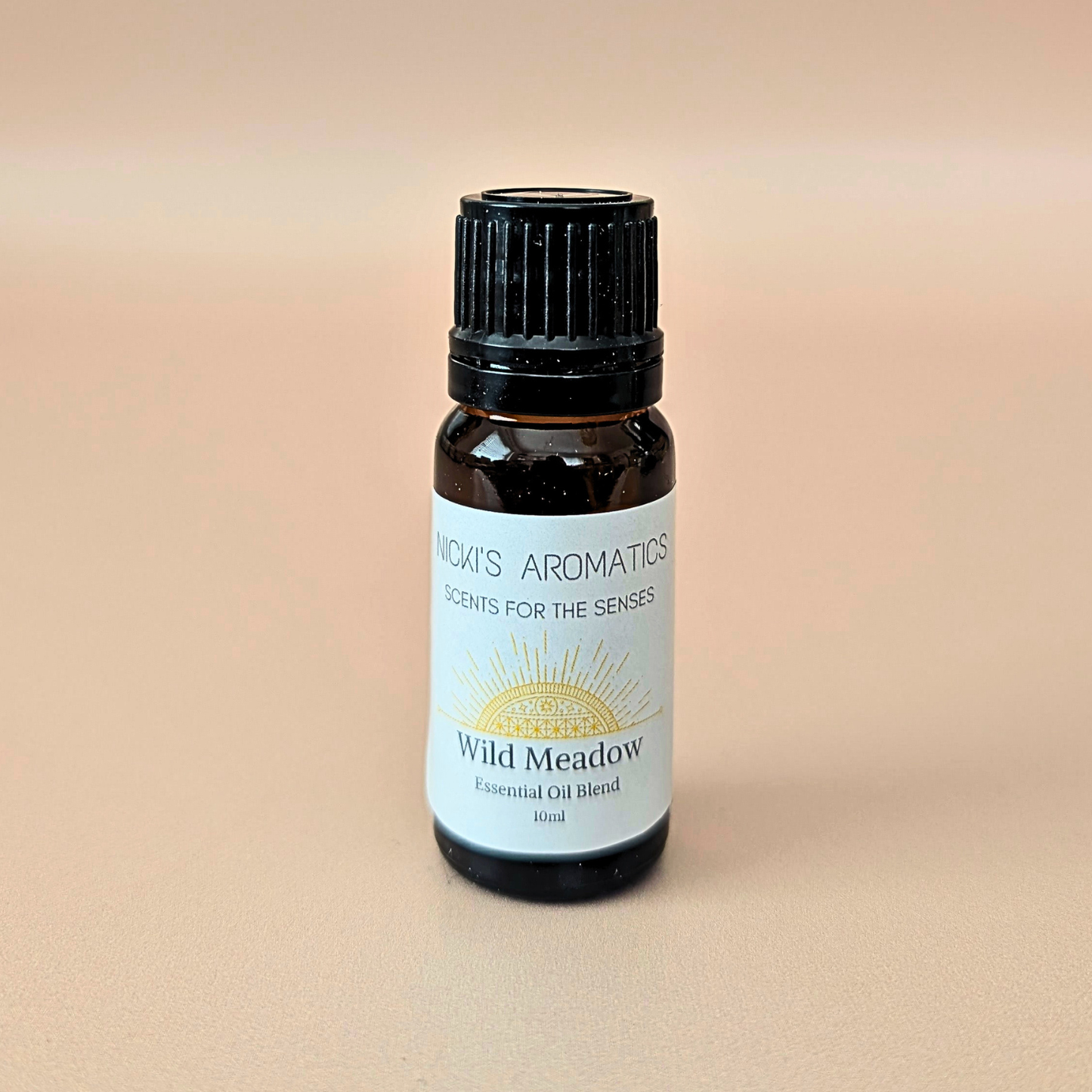 A small amber glass bottle of NICKI'S AROMATICS 'Wild Meadow' essential oil blend made with orange, ylang-ylang and cedarwood, with a black ribbed cap and a white label, set against a solid beige background.